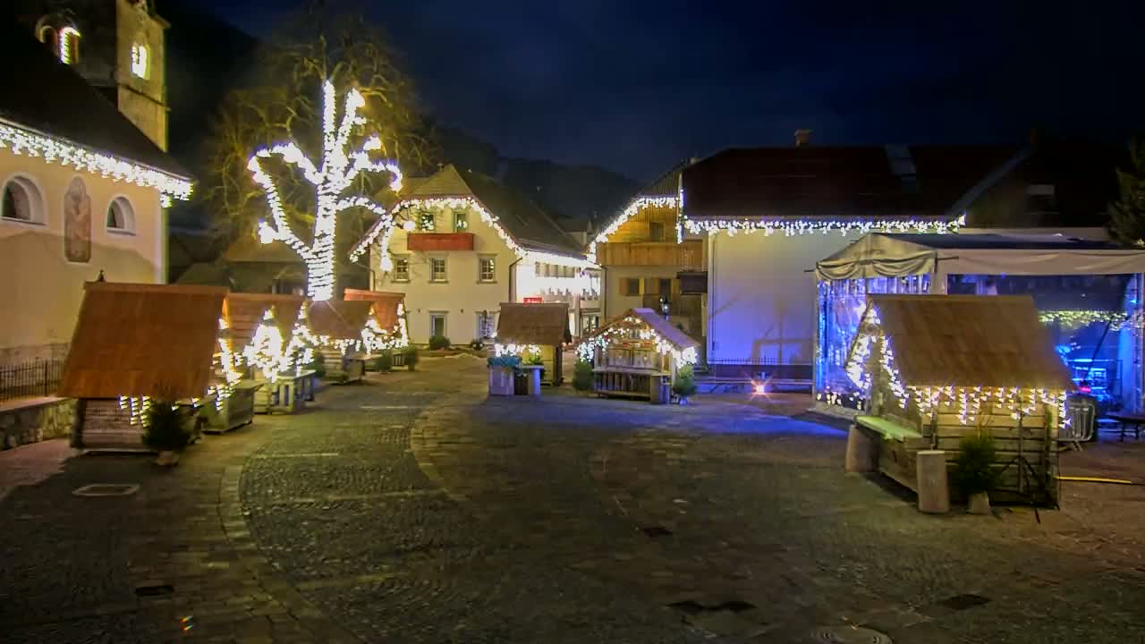 A nighttime scene shows a town square decorated with Christmas lights illuminating small wooden market stalls and a large tree, all set against a backdrop of buildings.