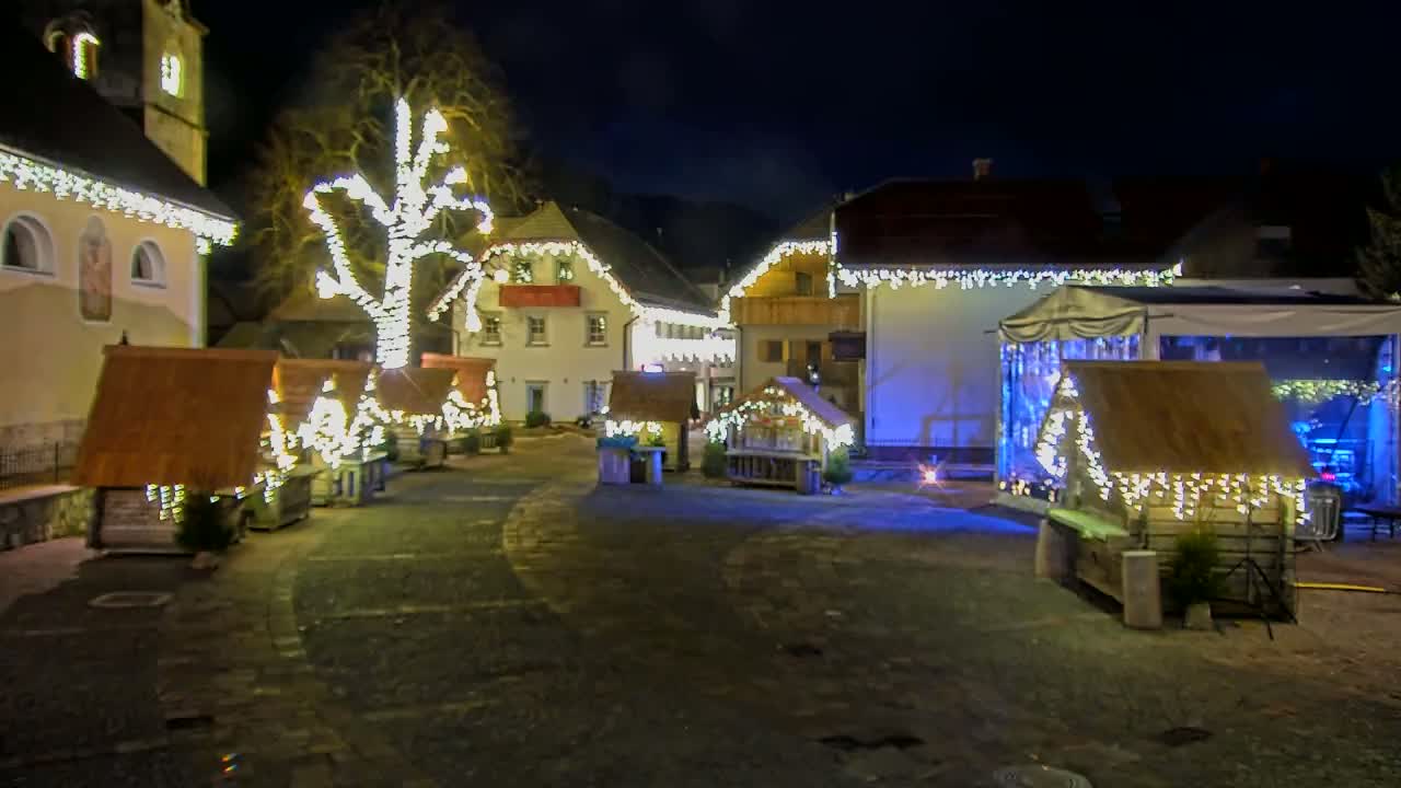 A nighttime scene of a town square decorated with Christmas lights illuminating small wooden structures and a large tree.