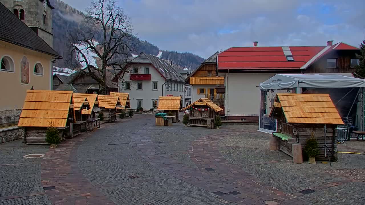 A cobblestone town square, surrounded by buildings and small wooden stalls, under a cloudy sky with snow-covered mountains in the background.
