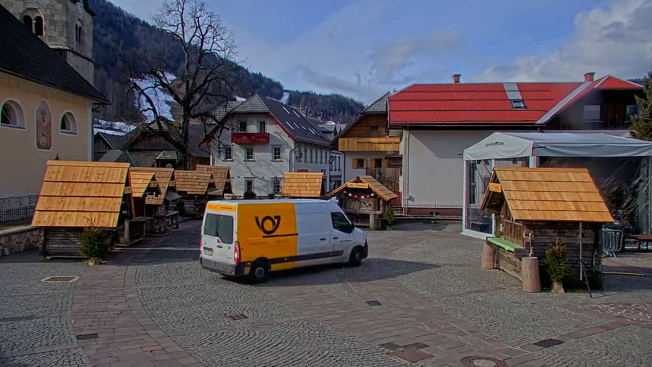 A white delivery van sits in a cobblestone town square surrounded by small wooden stalls and buildings, with snow-capped mountains in the background under a mostly sunny sky.