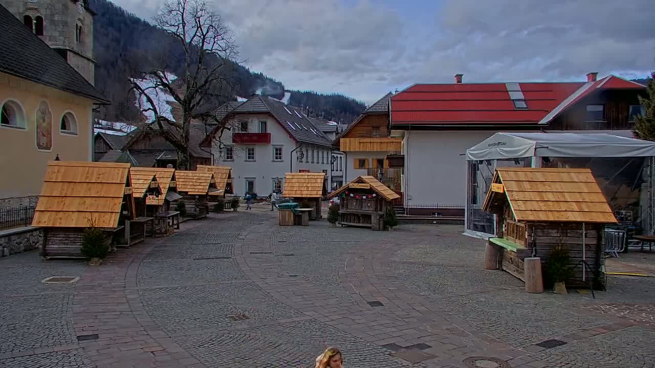 A cobblestone town square with several small wooden stalls, surrounded by buildings and a snow-covered mountain in the background under a cloudy sky.