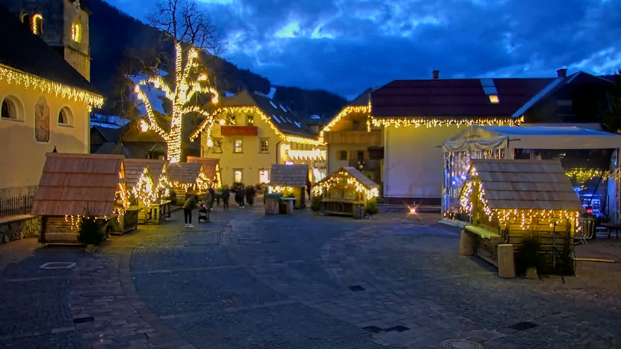 A town square at night, illuminated with string lights on buildings and small wooden market stalls, under a twilight sky.