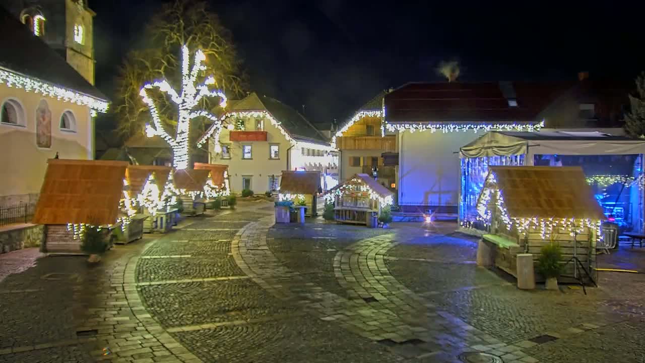 A cobblestone town square at night is illuminated by strings of lights adorning buildings and small wooden stalls.