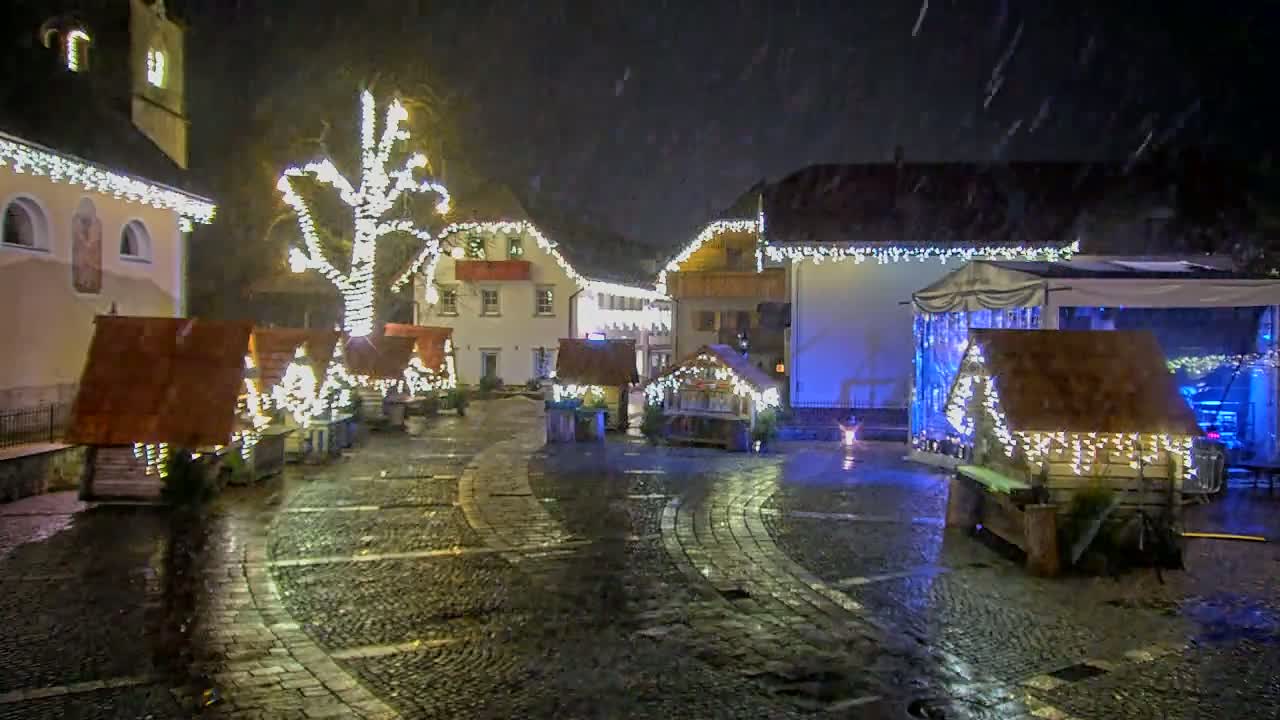 A snow-covered cobblestone town square at night is illuminated by Christmas lights strung on buildings and small wooden stalls.