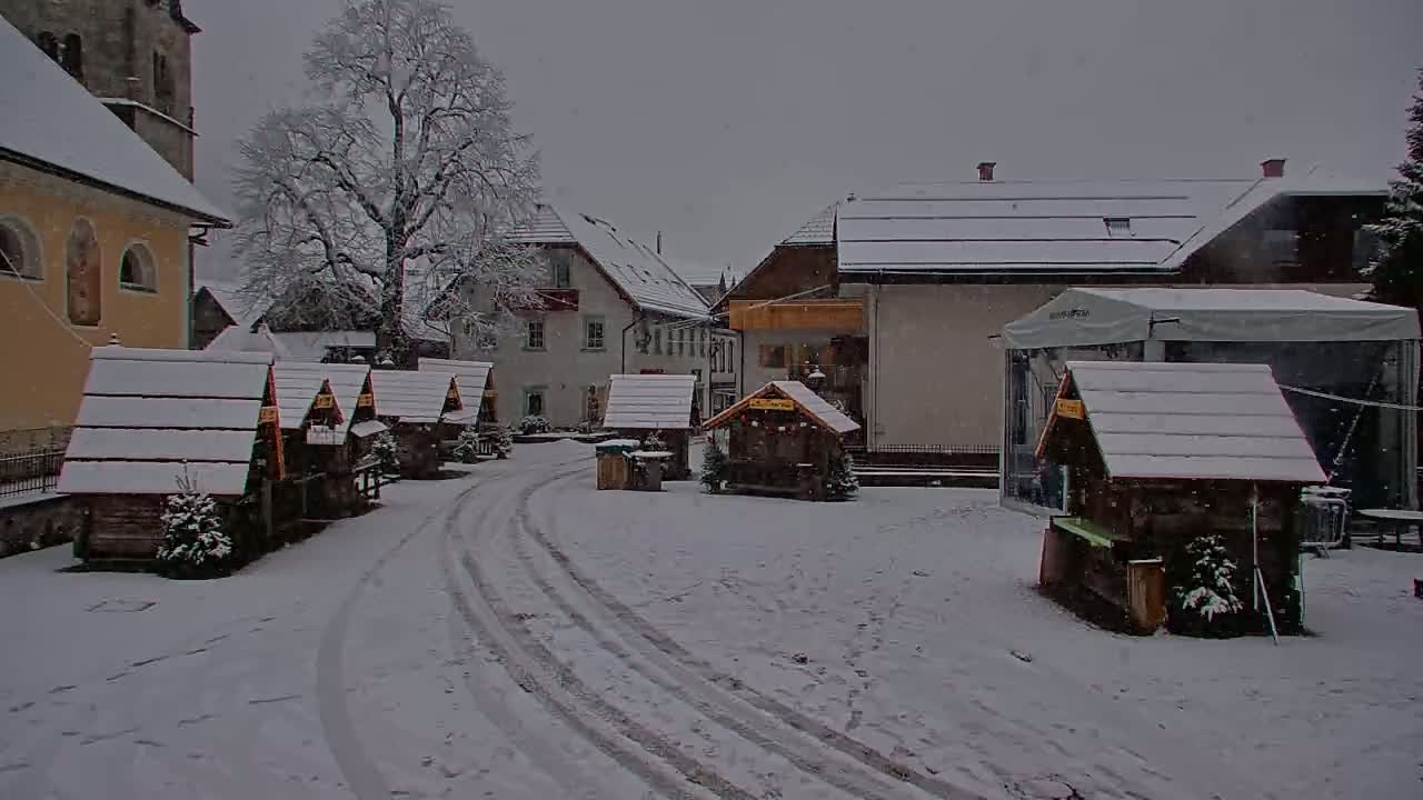 A snow-covered village square with several small wooden structures and buildings, under a light snowfall.