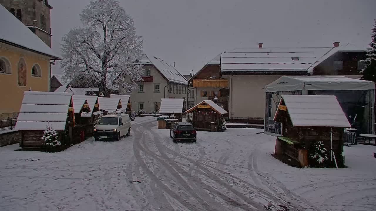 A snow-covered village square with several small wooden market stalls, a few cars, and buildings, under a light snowfall.