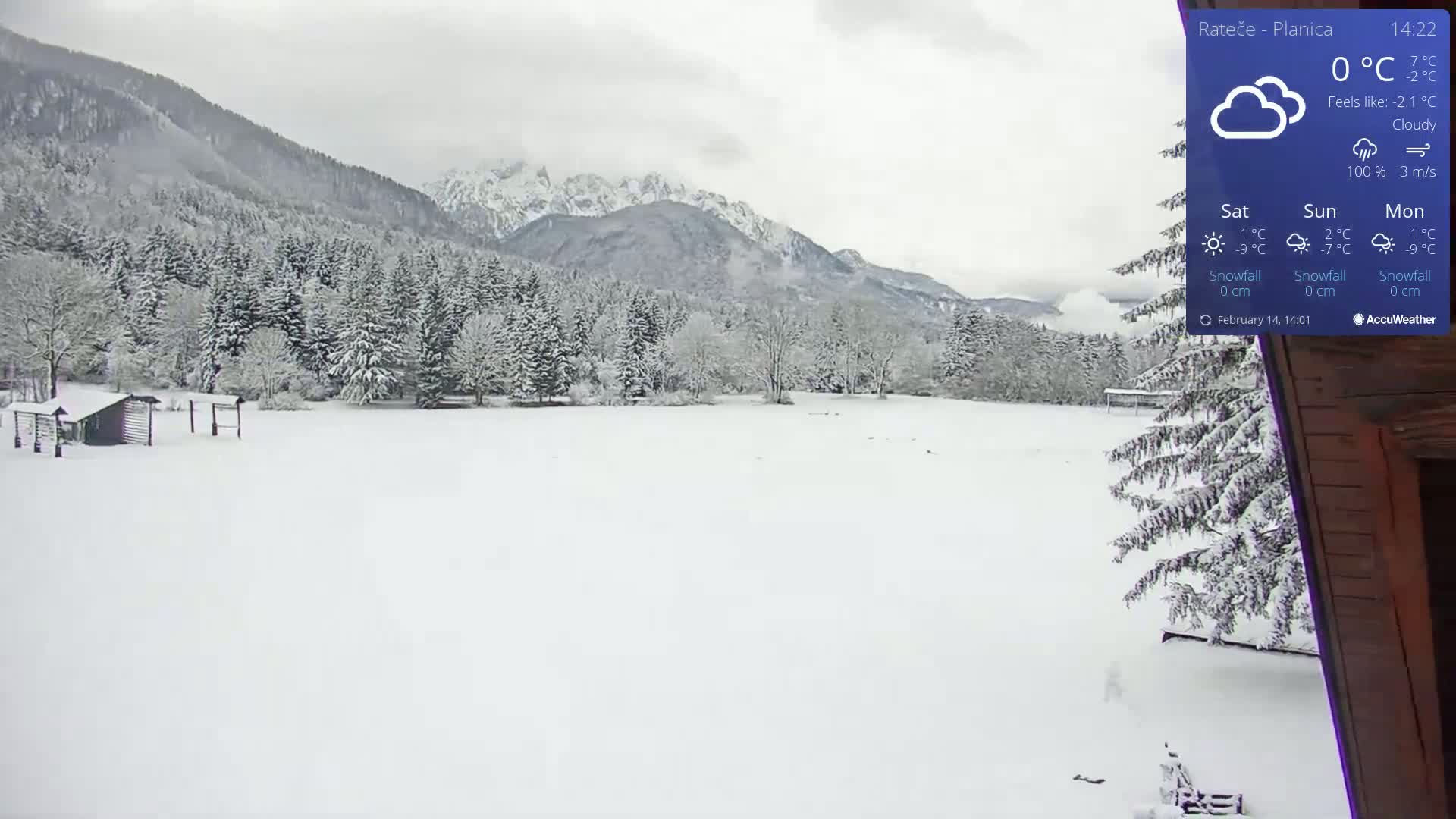 A snow-covered landscape with a small wooden building and snow-laden trees in the foreground, stretching to snow-capped mountains under a cloudy sky.