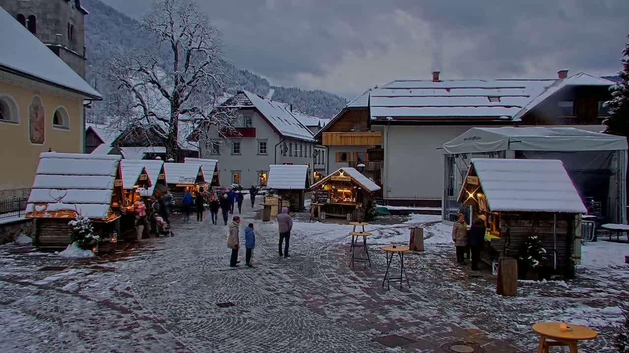 A snow-covered town square features several small wooden market stalls surrounded by people, with snow-covered buildings and a tree in the background under an overcast sky.