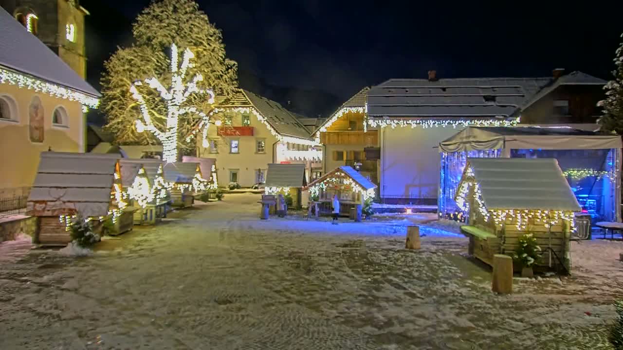 A snow-covered village square at night is illuminated by Christmas lights strung on buildings and small wooden huts.