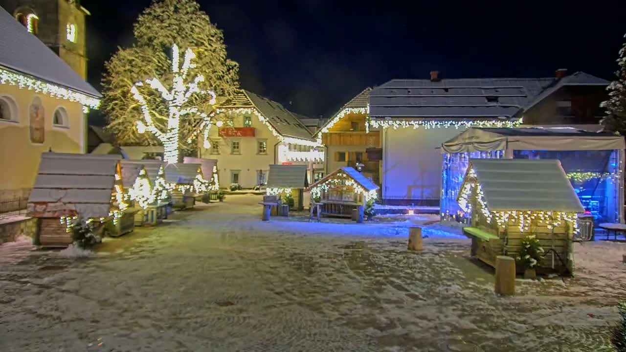 A snow-covered village square at night is illuminated by strings of lights adorning buildings and small wooden huts.