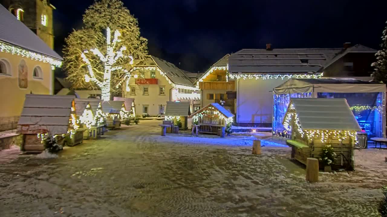 A snow-covered village square at night is illuminated by strings of lights adorning buildings and small wooden structures.