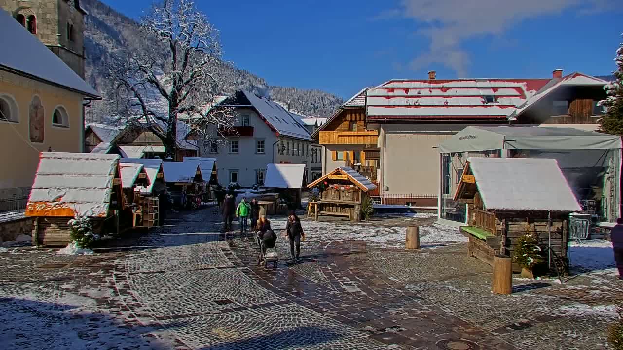 A snow-covered village square features several small wooden market stalls, a few pedestrians, and buildings with snow-laden roofs, all set against a backdrop of snow-dusted mountains under a sunny sky.