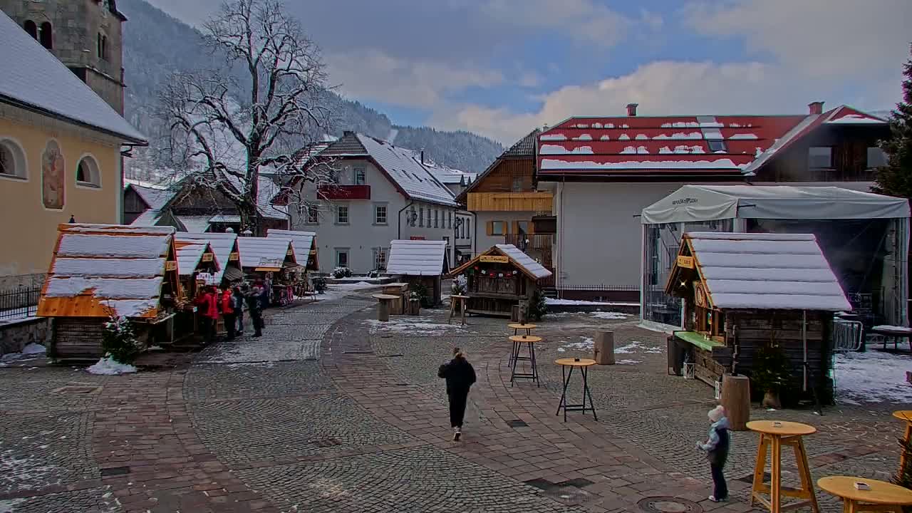 A snow-covered town square with small wooden market stalls, buildings, and a large tree under a partly cloudy sky.