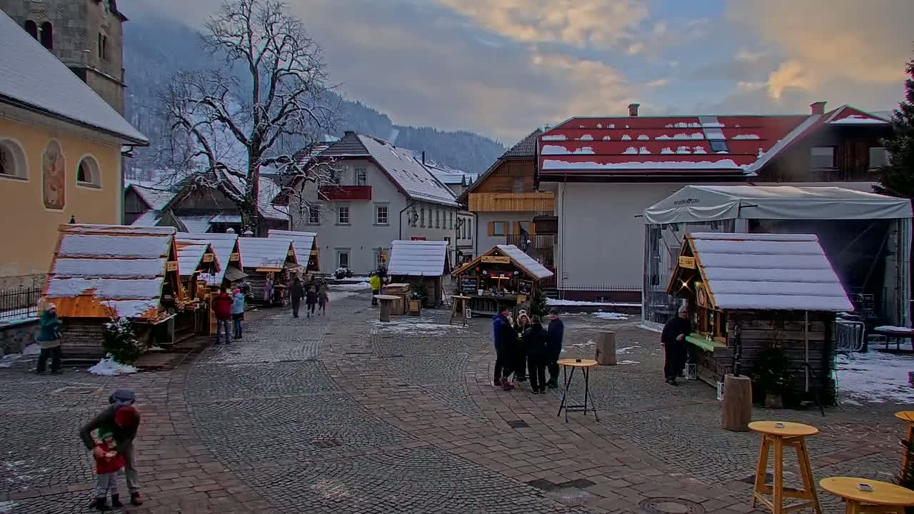 A snow-covered town square features several small wooden market stalls, surrounded by buildings and people, under a cloudy sky.