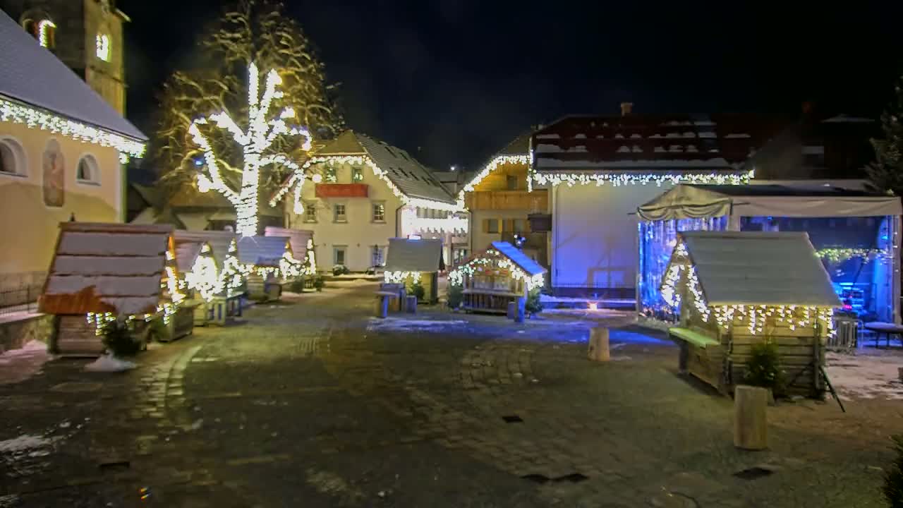 A snow-covered village square at night is illuminated by strings of lights on buildings and small wooden structures.