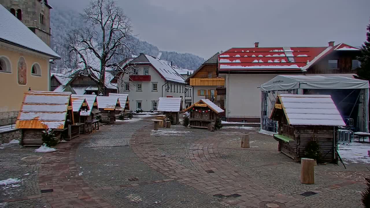 A snow-covered village square features several small wooden stalls, buildings with snow-dusted roofs, and a backdrop of snow-capped mountains under a cloudy sky.
