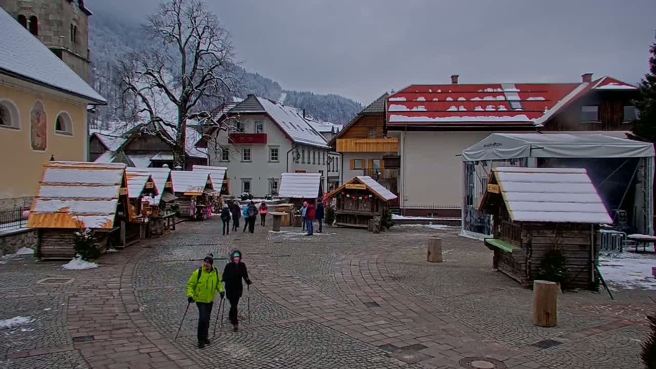 A snow-covered village square features several small wooden market stalls, a few people walking, and buildings with snow on their roofs, all set against a backdrop of snow-dusted mountains under an overcast sky.