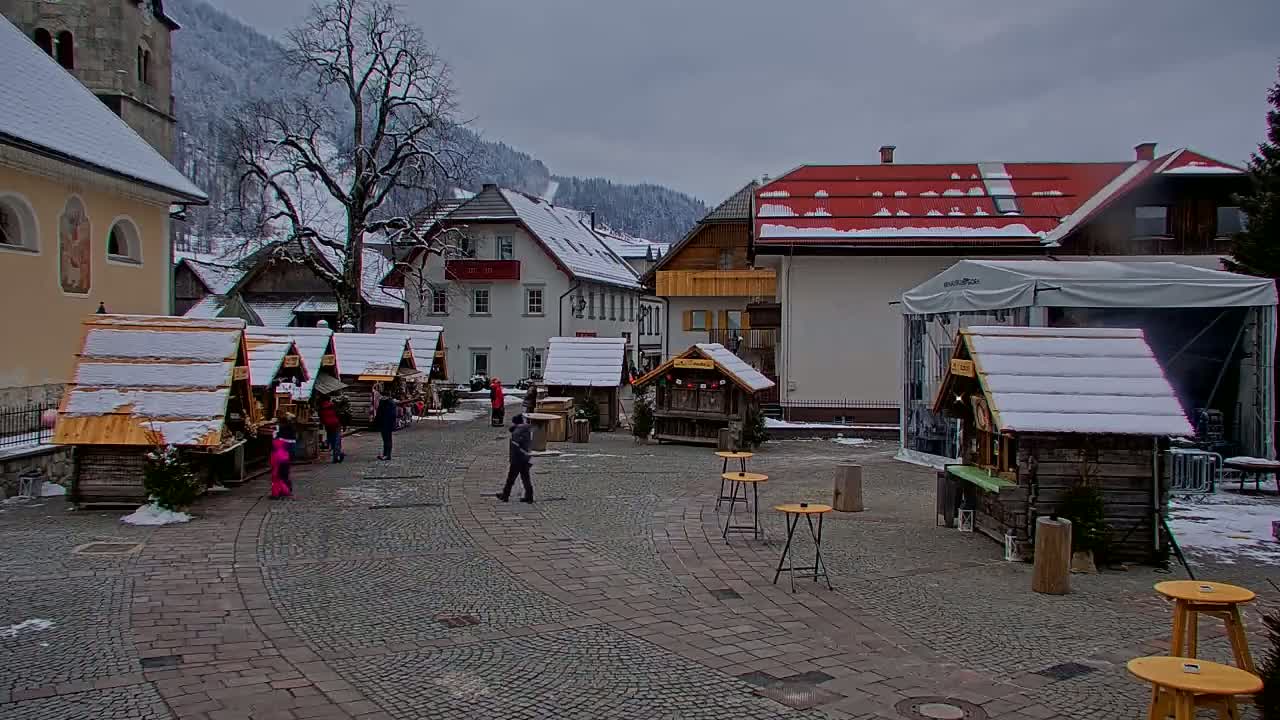 A snow-covered market square with wooden stalls and a few people walking around, situated in a village nestled in a snowy mountain valley under an overcast sky.