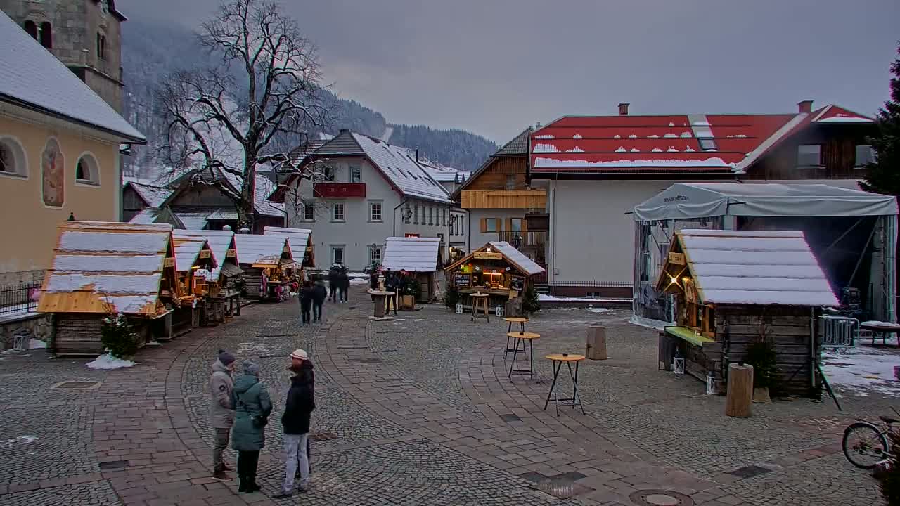 A snow-covered Christmas market occupies a cobblestone town square, surrounded by buildings and a backdrop of snow-dusted mountains under a cloudy sky.