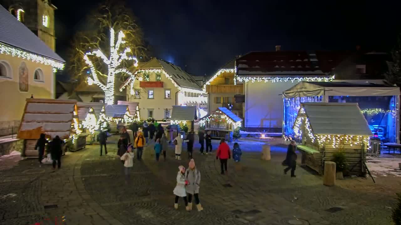 A nighttime scene of a town square with numerous small wooden structures decorated with lights, a large tree also lit up, and people strolling around.