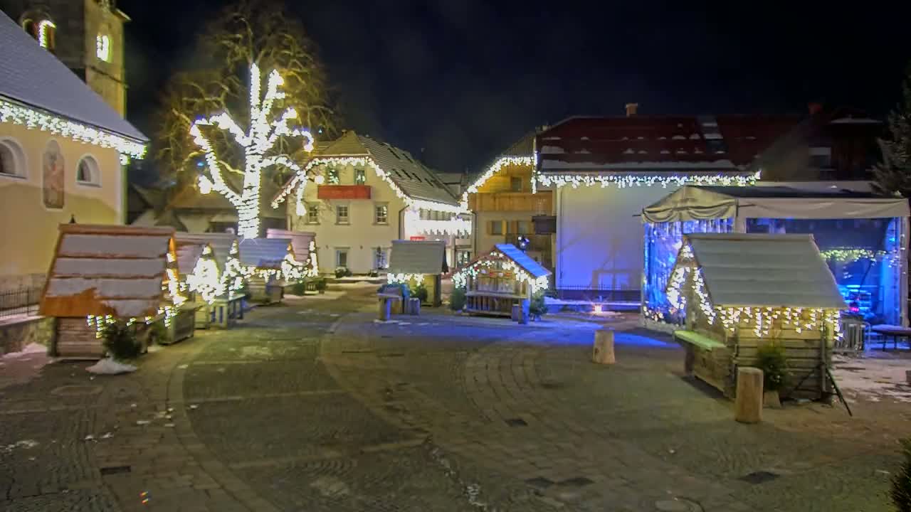 A snow-dusted village square at night is illuminated by strings of lights adorning buildings and small wooden huts.