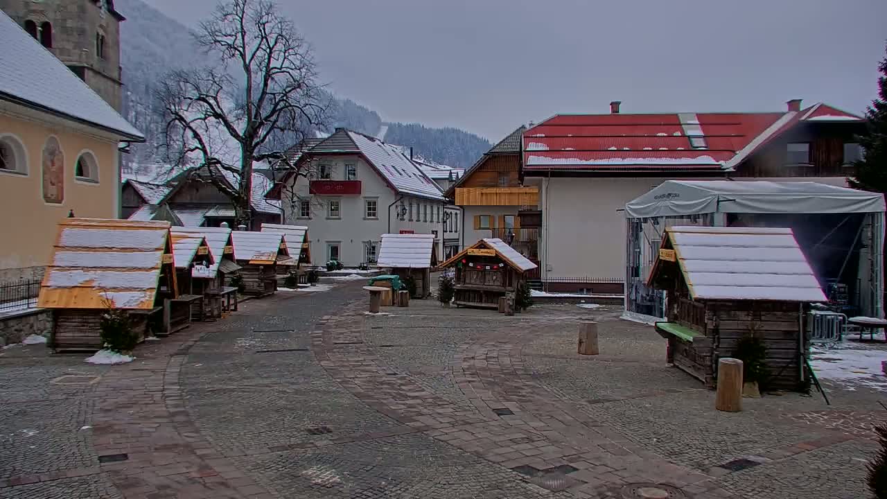 A snow-covered village square features several small wooden stalls and buildings, with a large bare tree in the background under an overcast sky.