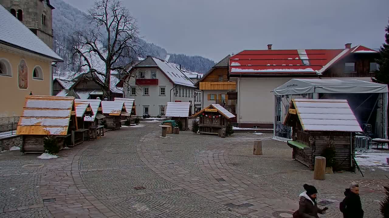 A snow-covered village square with several small wooden huts, buildings with red roofs, and a few people walking.