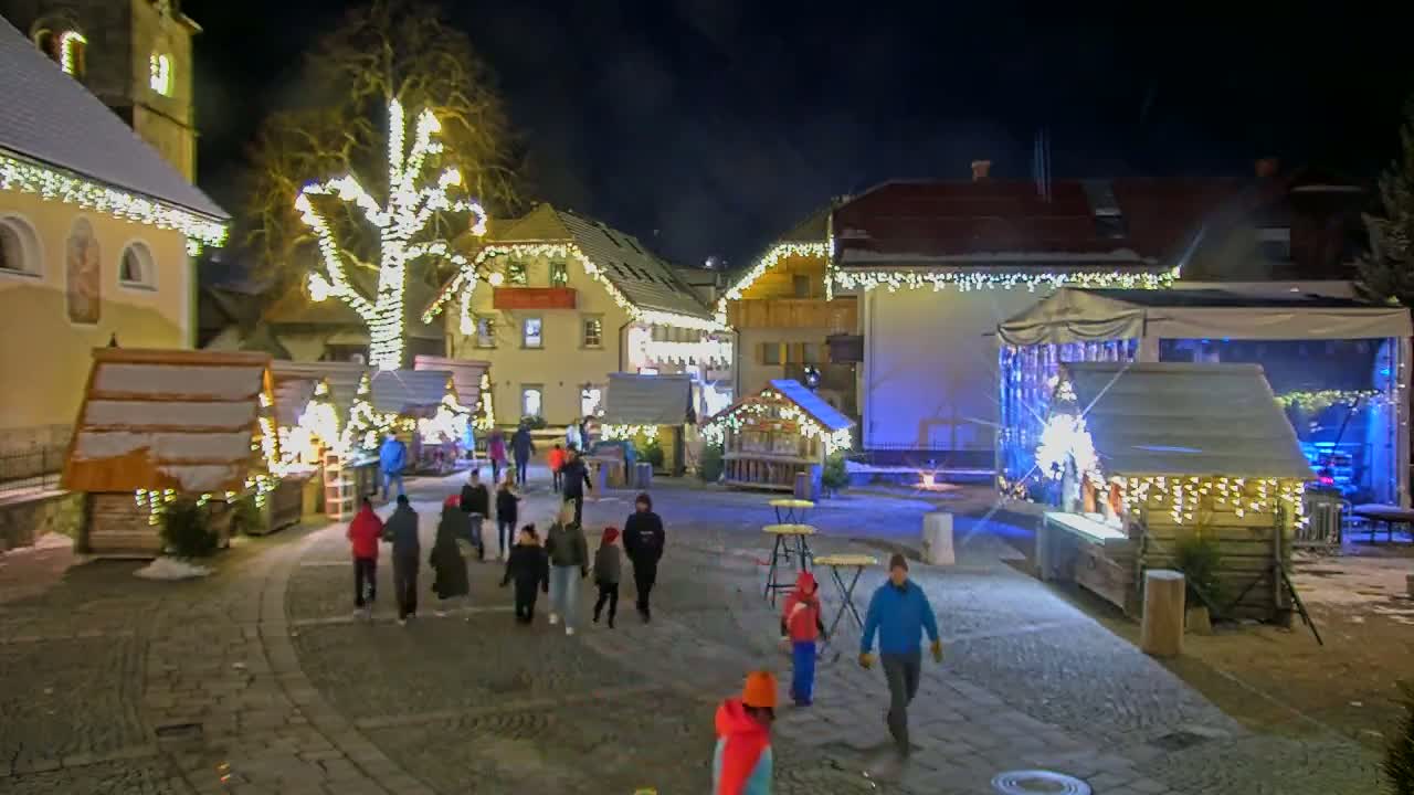 A nighttime scene of a snow-dusted Christmas market in a village square, illuminated by strings of lights on buildings and a central tree, with people walking among wooden stalls and tables.