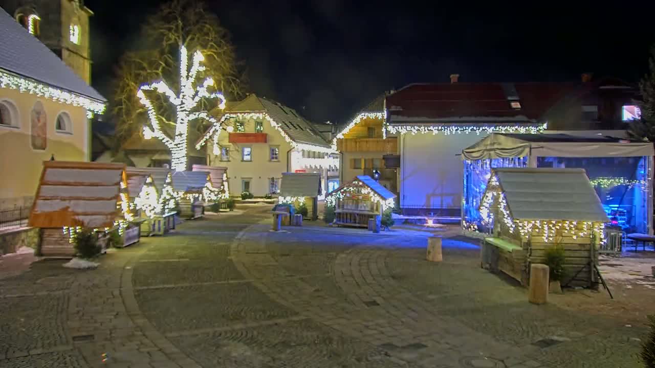 A snow-dusted town square at night is illuminated by strings of lights adorning buildings and small wooden stalls.