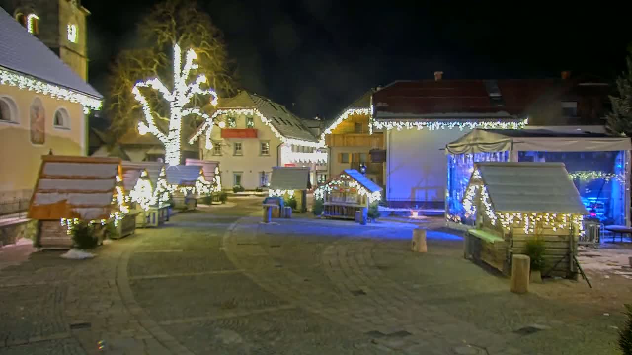A snow-dusted village square at night is illuminated by strings of warm white lights strung on buildings and small wooden market stalls.