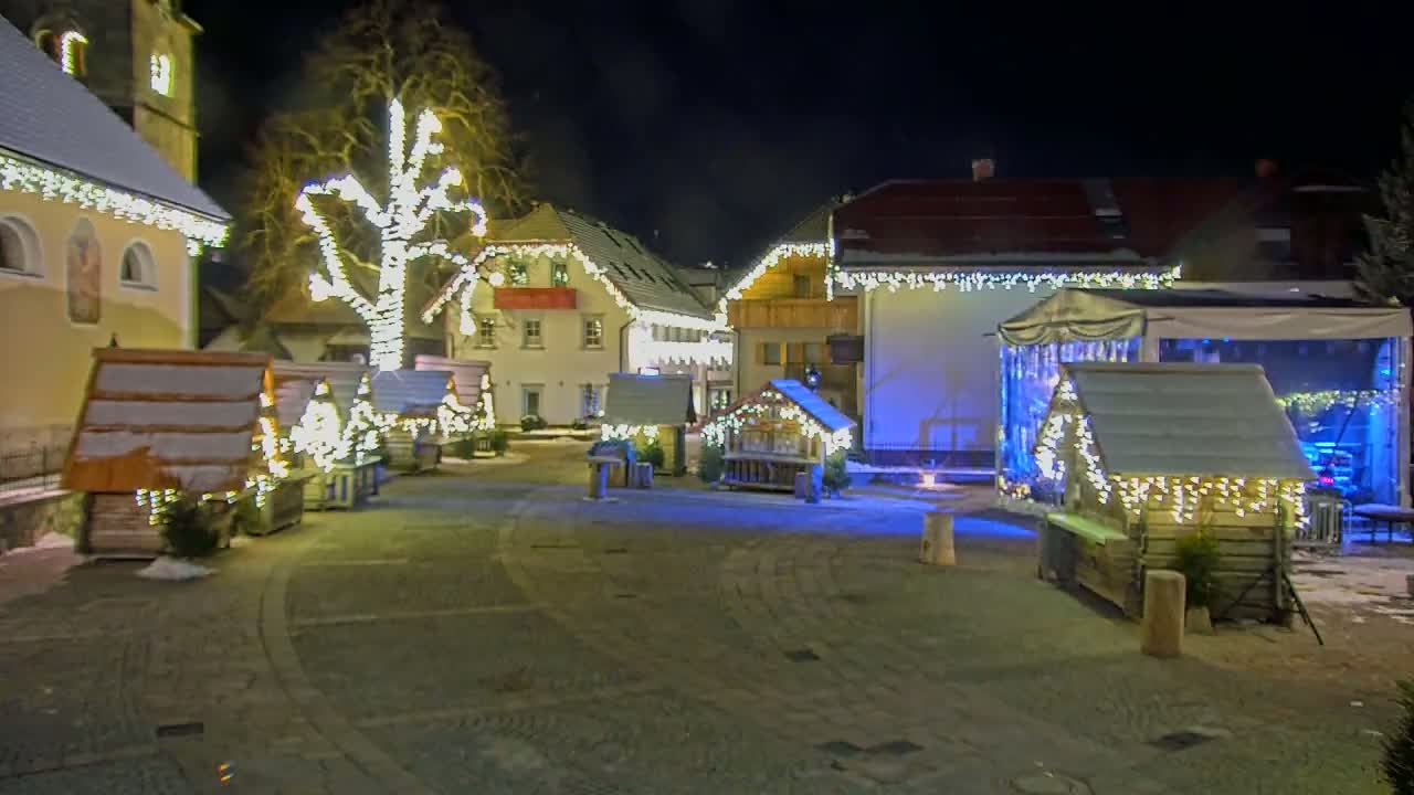A nighttime scene of a snow-dusted village square with small wooden huts decorated with lights, a large tree strung with lights, and buildings also adorned with lights.