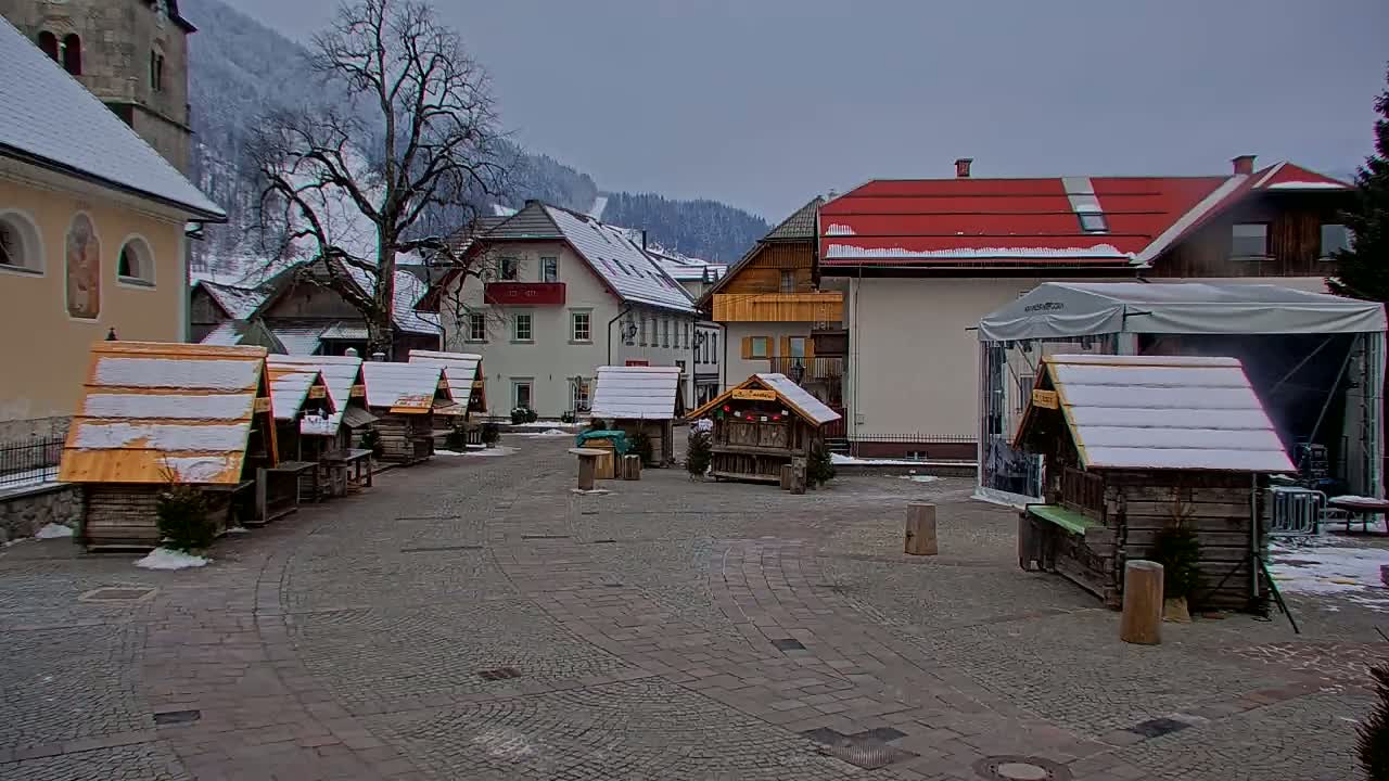 A snow-dusted village square features several small wooden market stalls and buildings, with a backdrop of snow-covered mountains under an overcast sky.