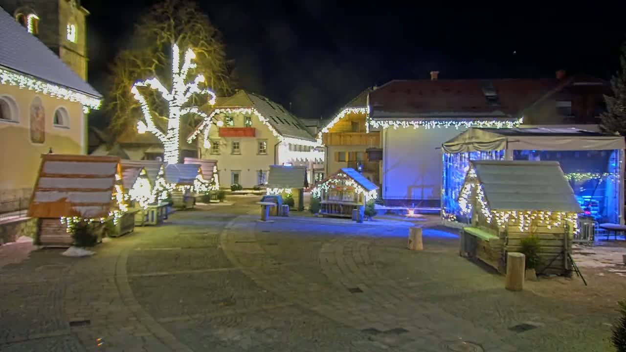 A nighttime scene of a town square decorated with Christmas lights and small wooden market stalls, with a few buildings illuminated in the background, on a slightly snowy ground.