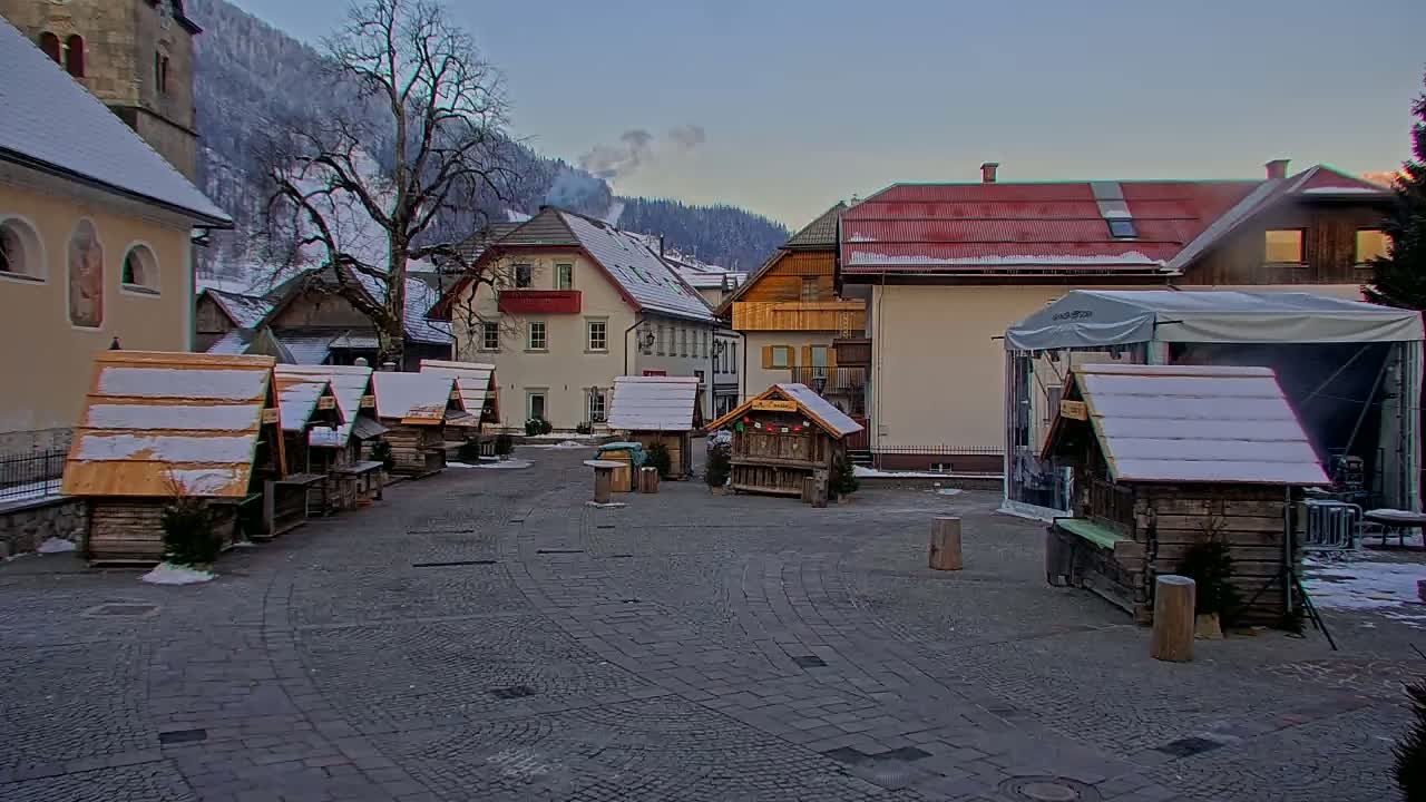 A snow-dusted village square features several small wooden market stalls, buildings with snow on their roofs, and a bare tree in the background against a backdrop of snow-covered mountains.
