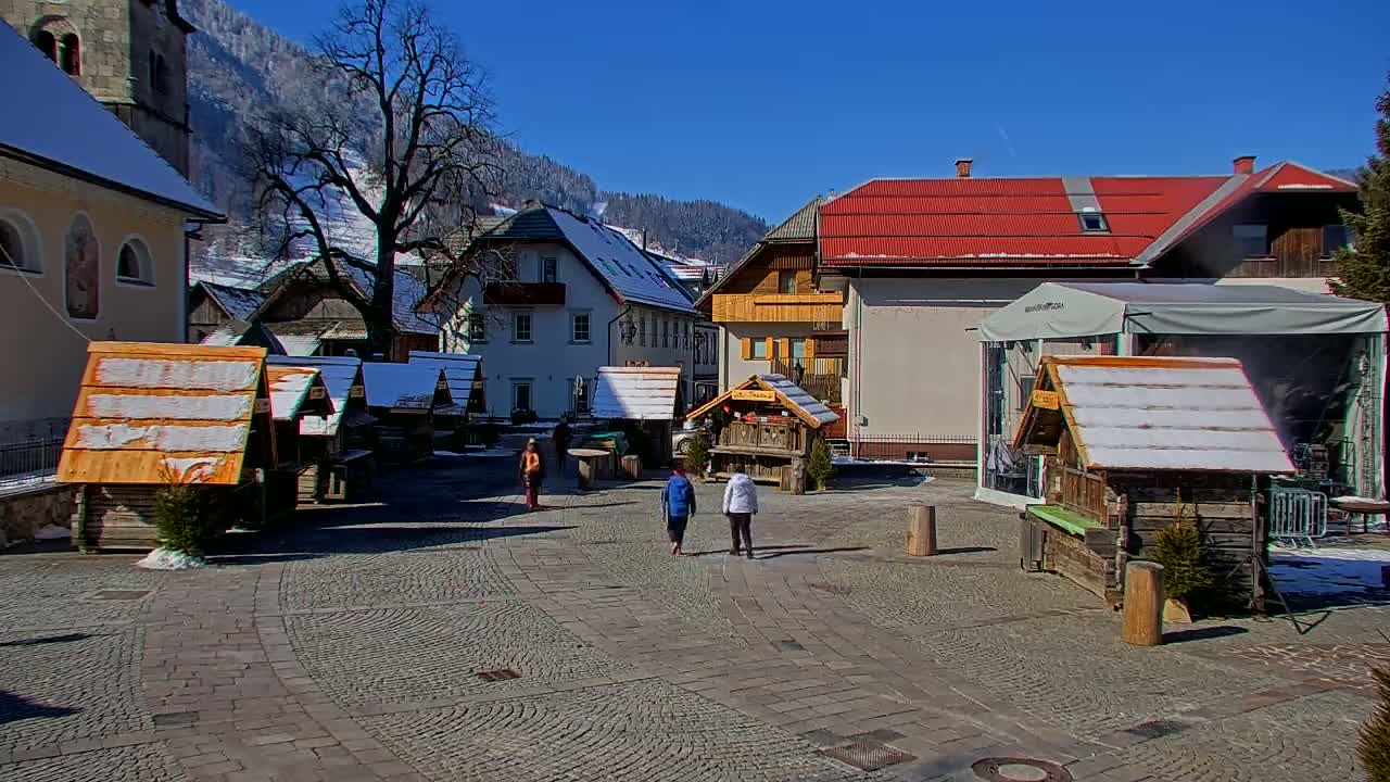 A cobblestone town square in a mountainous region, under a sunny sky, features several small wooden market stalls with snow on their roofs, and a few people walking by.