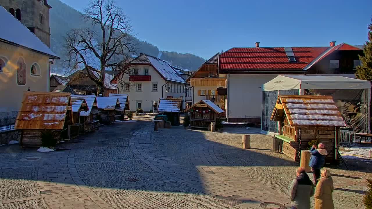 A cobblestone town square with several small wooden market stalls covered in snow, surrounded by buildings and a large tree, under a sunny winter sky.