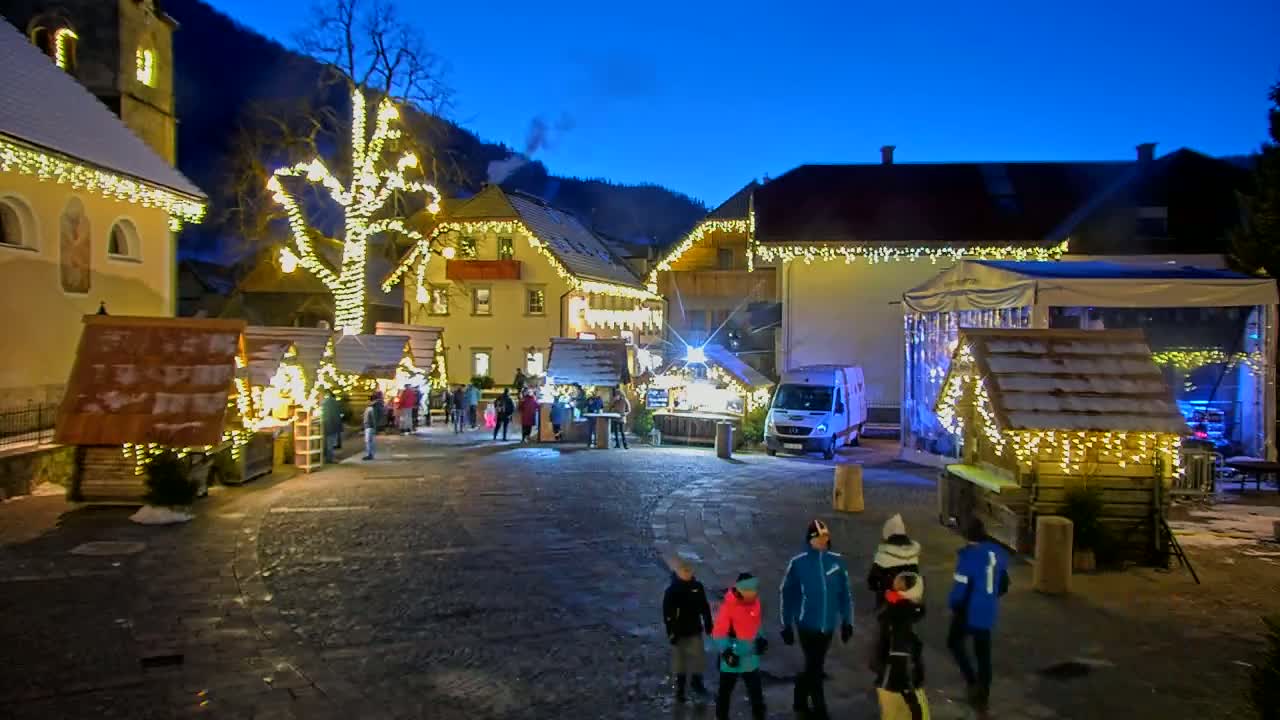 A nighttime outdoor Christmas market in a village, illuminated by string lights and featuring small wooden stalls, a large stage, and people strolling around.