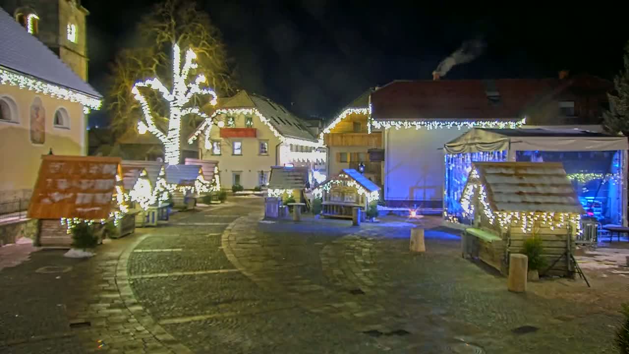 A nighttime scene of a village square decorated with Christmas lights, small wooden structures, and a large tree, with some snow on the ground.