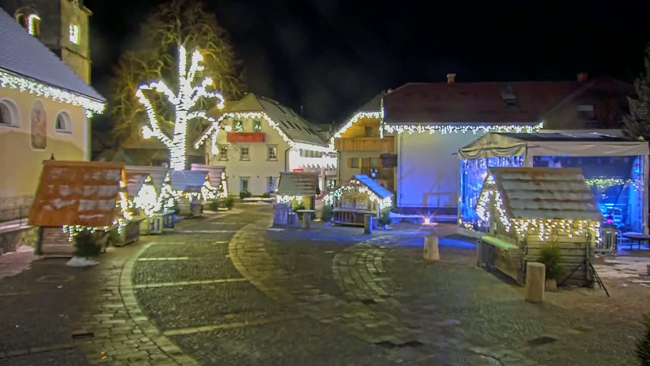 A cobblestone town square at night is decorated with Christmas lights strung on buildings and small wooden structures.