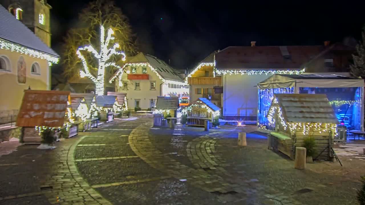 A cobblestone town square at night is decorated with small wooden huts and Christmas lights strung on buildings and trees.