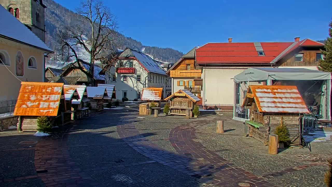 A sunny day in a snowy European village square shows several small wooden market stalls and buildings, with mountains in the background.