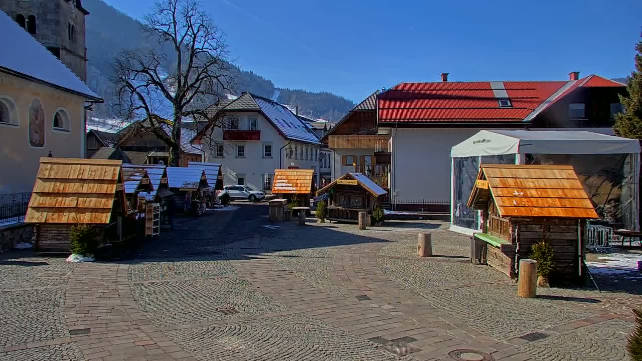 A cobblestone town square features several small wooden market stalls under a sunny, clear sky, with snow on the ground and surrounding buildings.