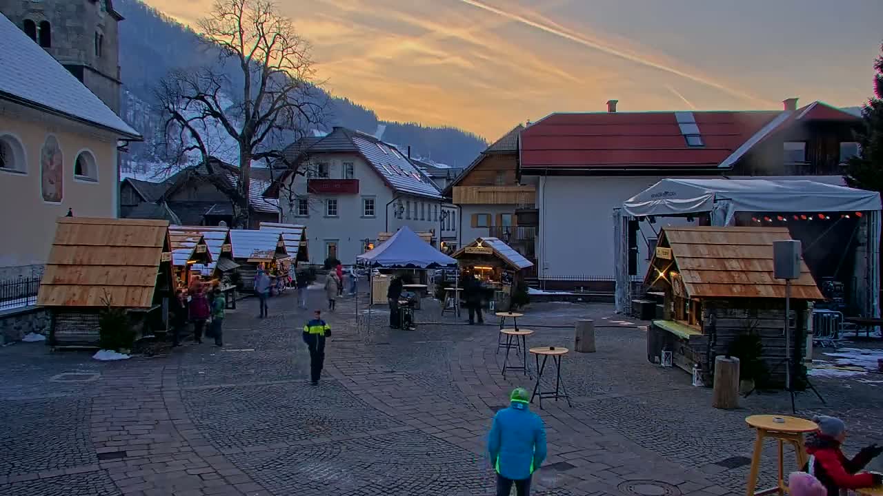 A cobblestone town square, under a twilight sky, features several wooden market stalls, a stage, and buildings with snow-covered roofs, set against a backdrop of snow-dusted mountains.