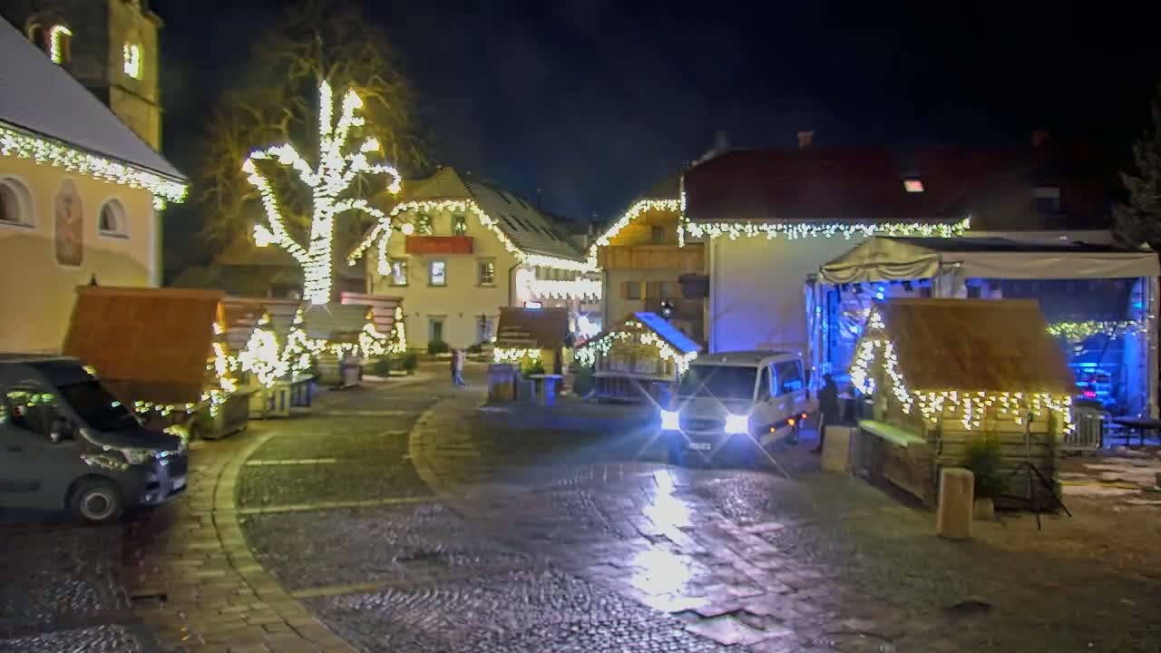 A nighttime scene of a village square adorned with Christmas lights, small wooden market stalls, and a stage, with a few vehicles parked nearby.