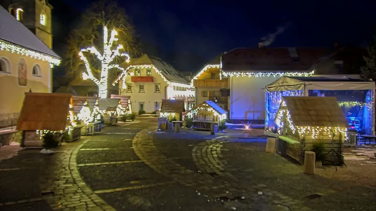 A cobblestone town square at night is decorated with small wooden huts and a large tree, all illuminated with strings of warm white lights; there is some snow on the ground.
