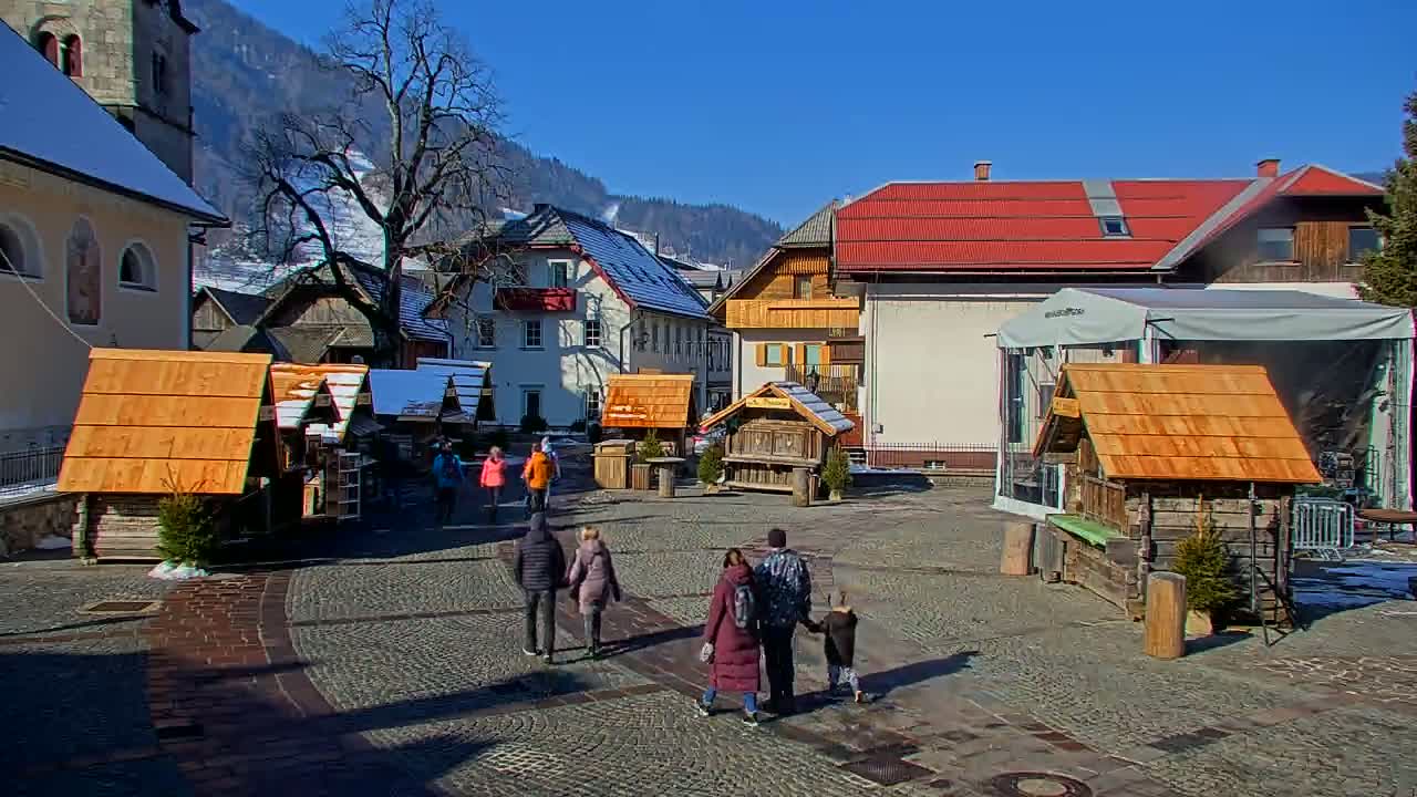 A cobblestone town square with several small wooden structures, a few people walking, and buildings with snow-covered roofs in the background under a sunny sky.