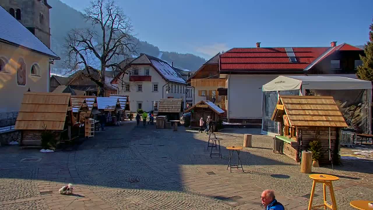 A sunny day in a cobblestone town square, featuring several small wooden market stalls, a few people, and snow-covered buildings against a backdrop of mountains.