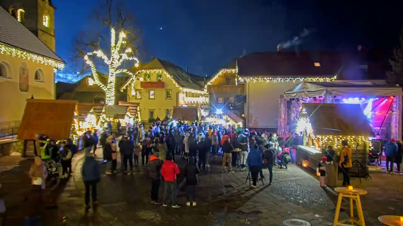 A nighttime outdoor Christmas market scene features many people gathered around market stalls and a stage in a town square decorated with lights.