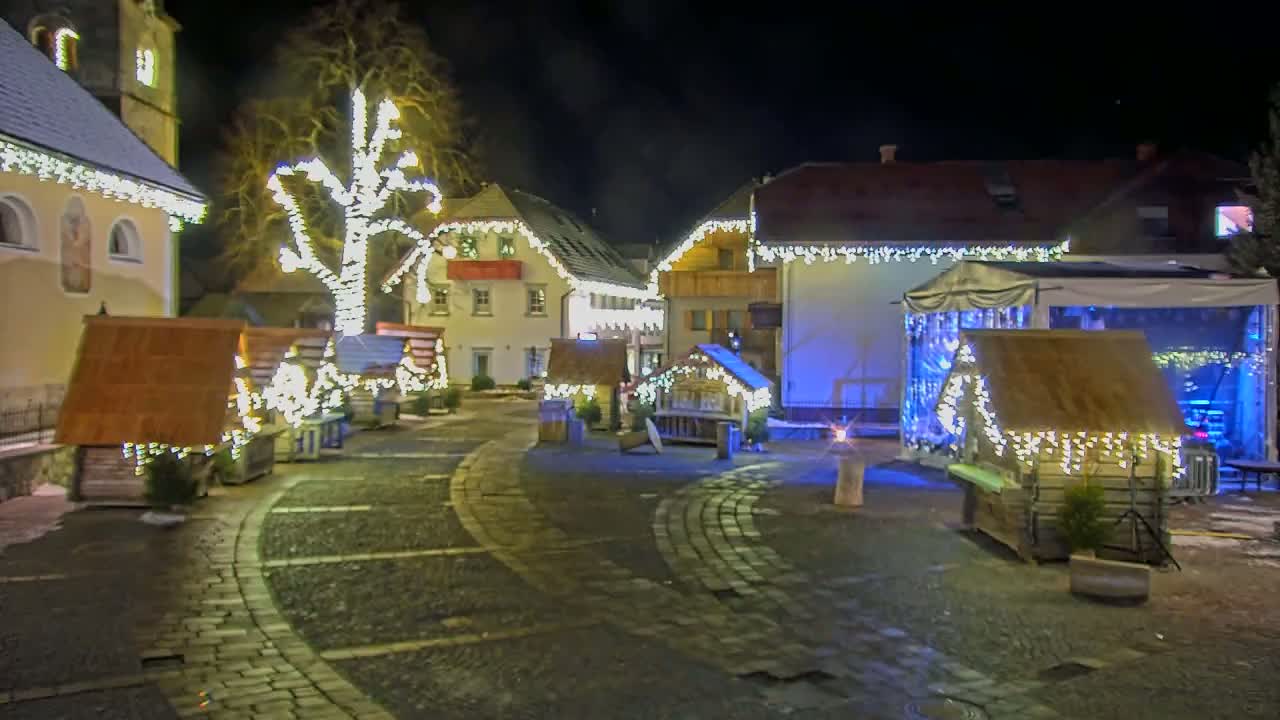 A nighttime scene of a town square decorated with Christmas lights, featuring small wooden structures and a large tree illuminated with lights.