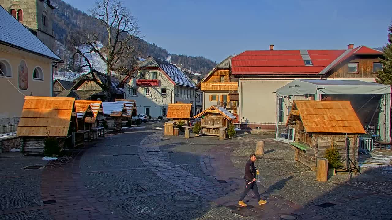 A cobblestone town square, under sunny skies, features several small wooden stalls and buildings, with a person walking in the foreground and snow-capped mountains in the background.